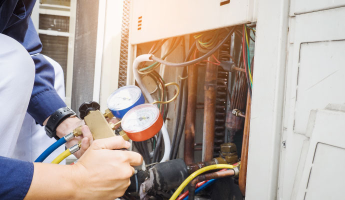 A person working on an outdoor HVAC unit, holding a manifold gauge to measure refrigerant pressures